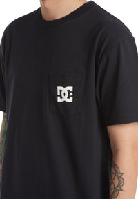 Černé bavlněné tričko s kapsou na hrudi a bílým logem DC. Materiál má hladkou texturu a standardní kulatý výstřih.
