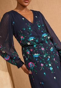 Rochie albastră navy cu broderie florală, având un decolteu în V, nasturi și mâneci bufante. Textura este ușoară, cu modele punctate.