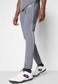 Grå joggebukser med hvite sidestriper og et brodert logo, laget av et elastisk stoff, kombinert med hvite joggesko.