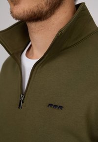 Pull-over en polaire zippé vert olive avec un col haut, comportant un motif brodé bleu marine. Texture lisse avec une coupe près du corps.