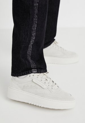 Baskets blanches à lacets portées avec un jean noir, montrées de côté sur un fond clair uni.