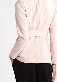 Světle růžový tailored blazer s páskem v pase, dlouhými rukávy a hladkým materiálem. Disponuje strukturovaným designem a diskrétními švy.