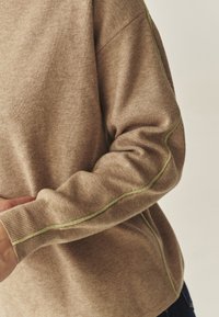 Hnedý pletený sveter s dlhými rukávmi, obsahujúci zelený akcentovaný šev pozdĺž švov. Mäkká textúra, voľný strih a rebrované manžety.