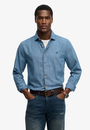 Mann trägt ein hellblaues Hemd mit hochgekrempelten Ärmeln, dunkelblaue Jeans, braunen Gürtel und Armbanduhr, Hände in den Taschen, blickt nach vorne.