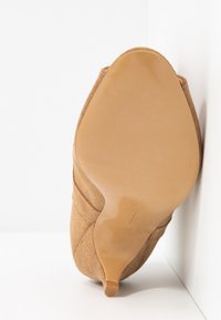 Brun ruskindshæl med en glat tan gummisål, buet form, subtile syninger langs siderne og et spidst tådesign.