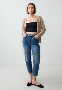 Hellbeige Strickjacke über einem schwarzen Cropped-Top, kombiniert mit hochsitztenden blauen Jeans. Glatte Textur, lockerer Schnitt, umgeschlagene Säume an den Jeans. Schwarze Ballerinas.