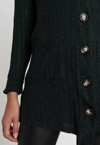 Tmavě zelený žebrovaný cardigan s uvolněným střihem, obsahující jednu náprsní kapsu a zlaté knoflíky pro zapínání. Měkká textura.
