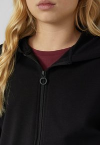 Zwarte rits-hoodie met een zachte textuur. Beschikt over een capuchon en een contrasterend bordeauxrood interior shirt dat zichtbaar is bij de halslijn. Zwarte ritshardware.