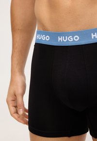 Boxer in cotone neri con un elastico azzurro chiaro che presenta la scritta "HUGO" in bianco. Tessuto liscio con una vestibilità aderente.