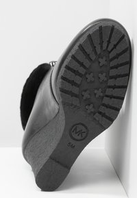 Bottine en cuir noir avec une semelle texturée présentant un motif en zigzag. Comprend un logo circulaire et un détail de la taille sur le côté.