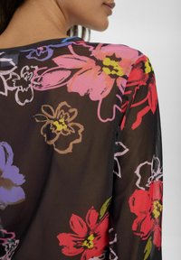 Blusa trasparente con stampa floreale su sfondo nero. Presenta grandi fiori rosa, rossi e viola con accenti verdi e un collo rotondo.