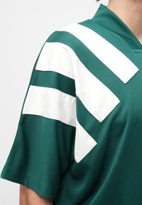 Žalia trumpomis rankovėmis marškinėliai su plačiomis baltomis chevron juostomis ant peties ir rankovės, dėvimi asmens, kurio figūra matoma nuo kaklo žemyn.