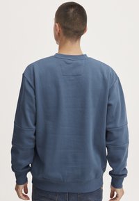Blauwe sweater met ronde hals, gestructureerde stof, verlaagde schouders en geribbelde manchetten. Heeft een klein achterpatch voor detail.