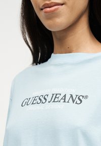 Γυναίκα που φοράει γαλάζιο πουκάμισο με το κείμενο «GUESS JEANS» και μικρότερο κείμενο τυπωμένα στο στήθος, ορατή από τους ώμους μέχρι το πηγούνι.