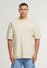 Beige oversized t-shirt made of cotton, featuring short sleeves, a round neckline, and a smooth texture. Worn with blue cargo shorts.