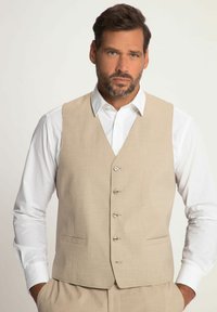 Beige Weste mit Reverskragen, Fronttaschen und fünf Knöpfen. Über einem weißen Hemd getragen. Glattes Gewebe mit feiner Textur.