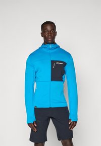 Kék cipzáras dzseki textúrált anyagból, fekete mellzsebbel és kapucnival. Sötét sort felett viselve, deréknál zsinórral.