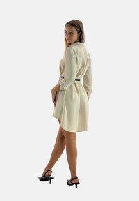 Vestito crema con colletto, maniche lunghe, vita stretta da una cintura nera e orlo svasato. Presenta una consistenza morbida e unDesign lungo fino alla caviglia.