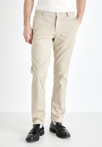 Beige bomullsbukser med slim fit, med glidelås og sidelommer, kombinert med svarte sko. Glatt tekstur, rent design.