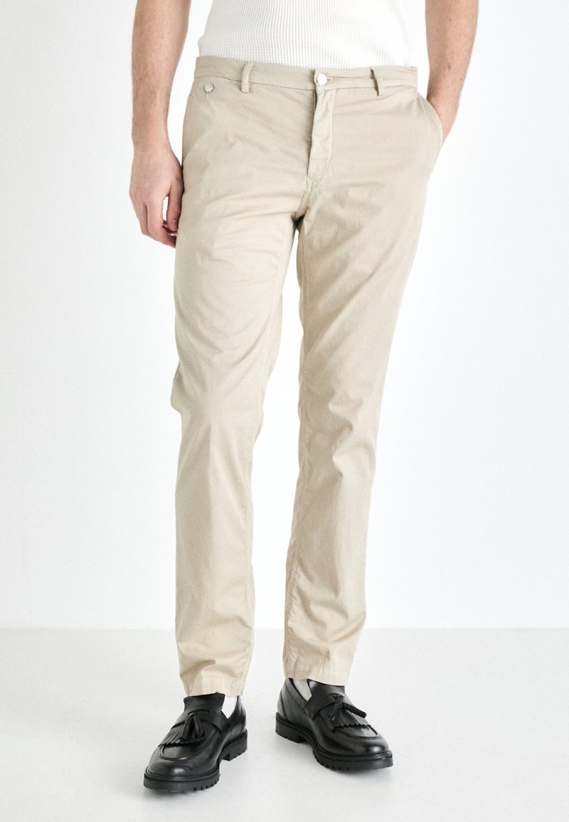 Beige bomullsbukser med slim fit, med glidelås og sidelommer, kombinert med svarte sko. Glatt tekstur, rent design.
