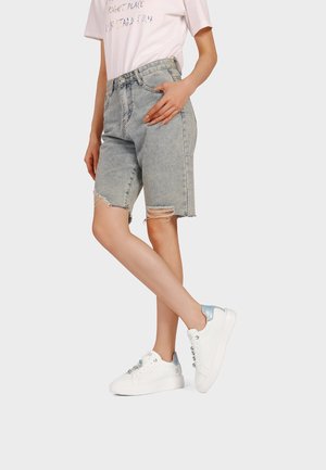 Persona che indossa pantaloncini di jeans chiari strappati fino al ginocchio, scarpe da ginnastica bianche con tacchi argentati e una maglietta rosa pallido a maniche corte con testo.