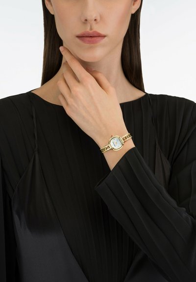 Montre dorée avec un cadran blanc rond, mettant en valeur un accent rouge. Portée sur un poignet fin contre un tissu noir plissé.