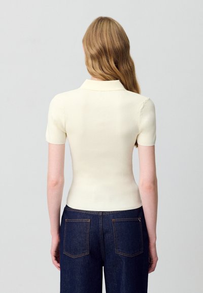 Camisa polo de canalé crema con manga corta, corte ajustado y collar estándar, combinada con jeans de denim azul oscuro con bolsillos traseros.