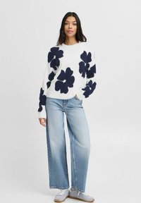 Weißer Pullover mit großen navyblauen Blumenmustern, kombiniert mit hellblauen, weiten Jeans. Der Pullover hat gerippte Bündchen und einen runden Ausschnitt.