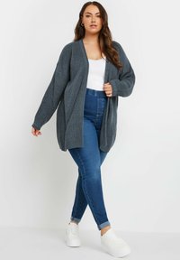 Cardigan grigio a coste con maniche lunghe, design aperto davanti, abbinato a jeans blu a vita alta e sneakers bianche. Dettaglio della trama liscia visibile.