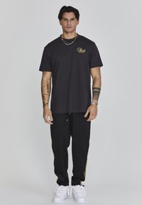 Camiseta negra de manga corta con logo dorado y pantalones de chándal negros con franjas doradas a los lados. Zapatillas blancas completan el atuendo.