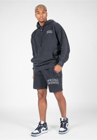 Sudadera negra con capucha y bolsillo frontal, logo "WRONG FRIENDS". Pantalones cortos negros a juego con detalle del logo. Llevado con zapatillas negras y blancas.