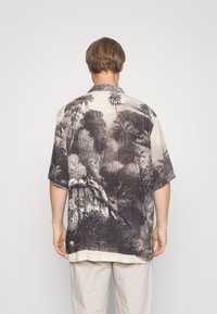 Uomo che indossa una camicia a maniche corte con stampa tropicale in bianco e nero, in piedi con la schiena rivolta verso uno sfondo chiaro e neutro.