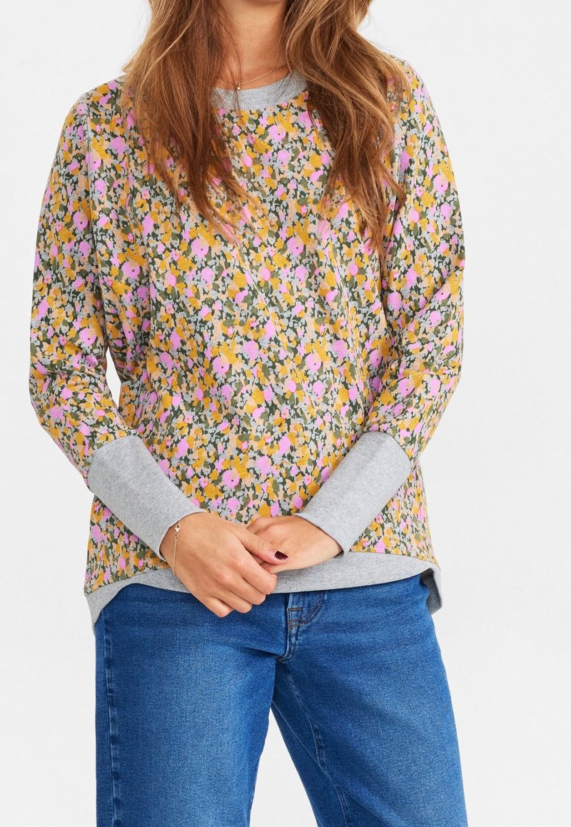 Femme portant un pull à manches longues à fleurs avec des poignets gris et un jean bleu, les mains légèrement jointes devant.