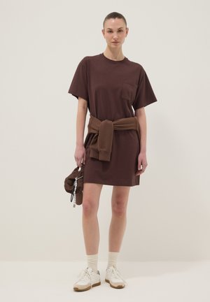 Donna che indossa un vestito marrone a maniche corte con una felpa legata in vita, tiene un berretto marrone, calzini bianchi e scarpe da ginnastica bianche.