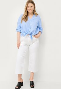 Blouse en lin bleu clair avec un détail de nœud à la taille, assortie à un pantalon blanc à jambes larges et des mules à talons ouverts noires.
