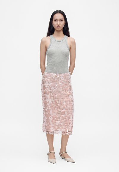 Jeune femme aux longs cheveux foncés porte un haut sans manches gris, une jupe midi rose à sequins et des ballerines pointues beiges, debout les mains derrière le dos.