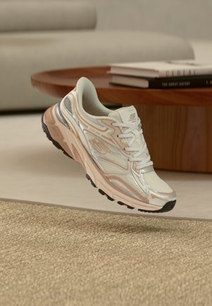 Scarpa da ginnastica bianca e beige con cuciture dettagliate, pannelli in mesh e suola spessa, sospesa sopra un tappeto beige intrecciato all'interno.