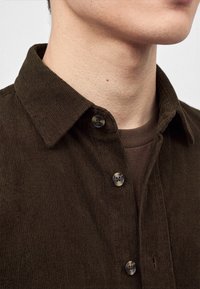 Gros plan d'une personne portant une chemise en velours côtelé marron foncé boutonnée sur un t-shirt marron col rond.
