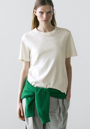 Vrouw die een eenvoudig wit T-shirt draagt, grijze broek en een groene trui om haar middel geknoopt, staand tegen een lichte achtergrond.