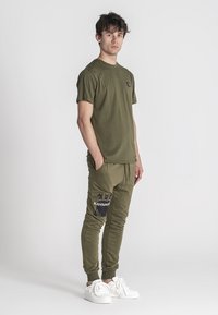 Camiseta de algodón verde oliva con cuello redondo; pantalones de chándal verdes con cinturilla elástica y acentos estampados. Zapatillas blancas completan el atuendo.