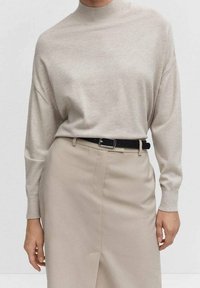 Pull en maille beige à col montant, épaules tombantes et manches longues, associé à une jupe taille haute beige clair et une ceinture noire.