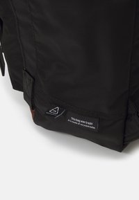 Schwarze Tasche aus recyceltem Nylon mit strukturierter Oberfläche, mit einer braunen Akzentfarbe und einem Etikett, auf dem steht: "Diese Tasche war Müll! Sie besteht aus recyceltem Nylon."