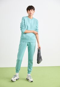 Lichtblauwe Nike sweatshirt en bijpassende joggers, beide gemaakt van zachte stof, met witte logo-accenten. Model draagt grijze sneakers en houdt een tas vast.