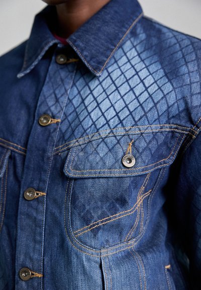 Denim jacket featuring a diamond pattern on the left side, blue fabric, gold stitching, and metal buttons with a pocket on the chest.