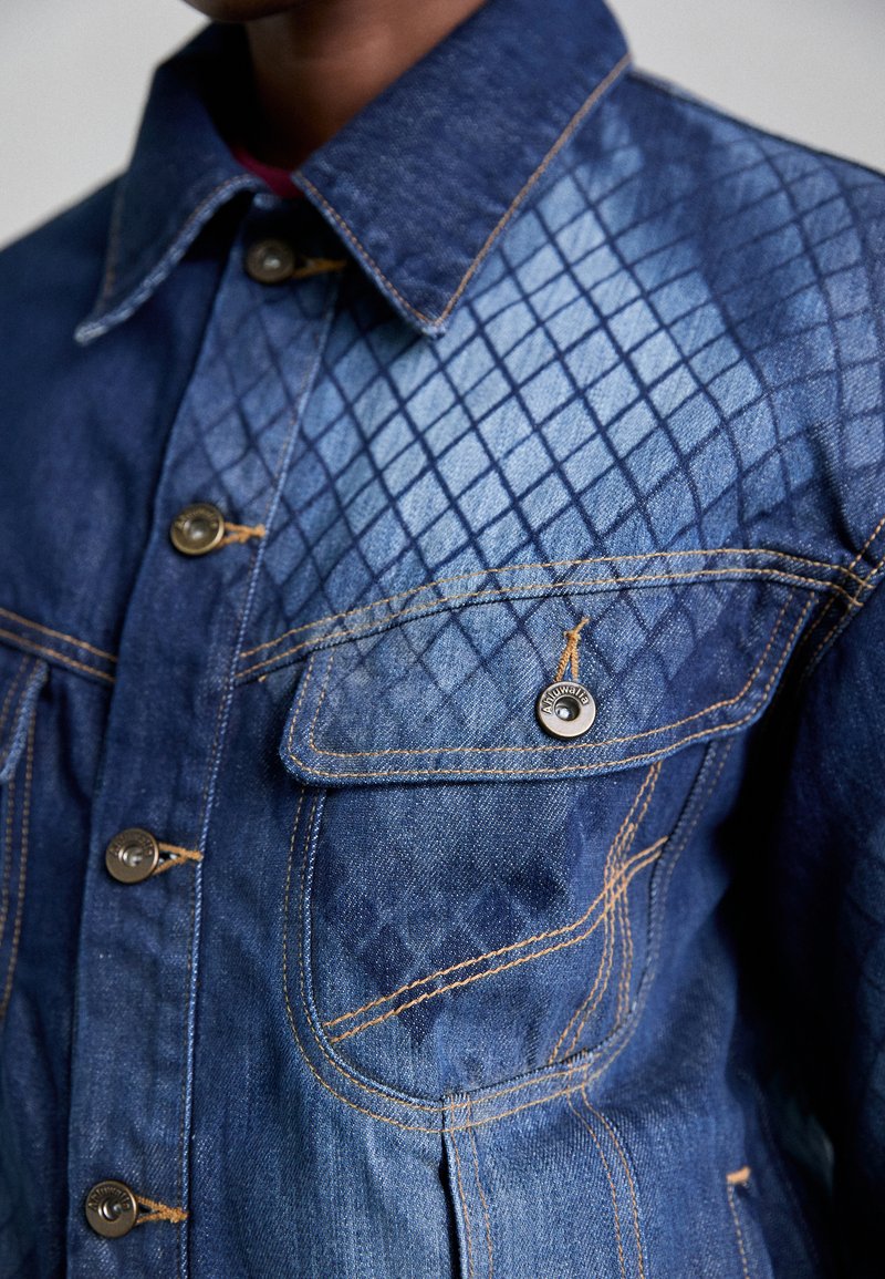 Denim jacket met een ruitpatroon aan de linkerkant, blauwe stof, gouden stiksel, en metalen knopen met een zak op de borst.