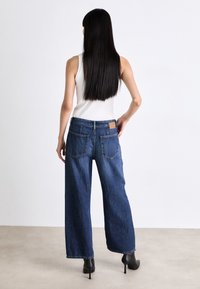 Weite Blue-Jeans aus Denim mit hohem Bund, aufgesetzten Gesäßtaschen und einem hellbraunen Lederlabel am Bund. Der Stoff hat eine weiche Textur.