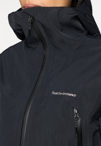 Μαύρο αδιάβροχο μπουφάν με ψηλό γιακά, μπροστινό φερμουάρ και λεπτομέρεια λογότυπου. Λείο υφή, μίνιμαλ σχεδίαση και δύο τσέπες με φερμουάρ.