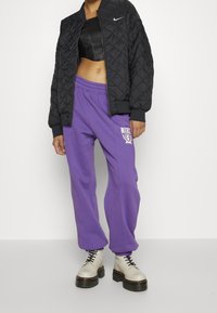 Černá prošívaná bomber bunda, krátký černý top, fialové tepláky s logem a krémové platformní boty s černými tkaničkami.