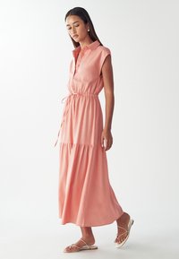 Długa różowa sukienka z kołnierzykiem, krótkimi rękawami, talią ze sznurkiem i falbanianym dołem. Gładki materiał o miękkiej teksturze.