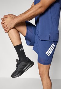 Μπλε αθλητικό σορτς με λευκές ρίγες, μαύρα αθλητικά παπούτσια και μαύρες κάλτσες. Δείχνει μια σκυφτή θέση ποδιού και λεπτομέρειες υφής υφάσματος.